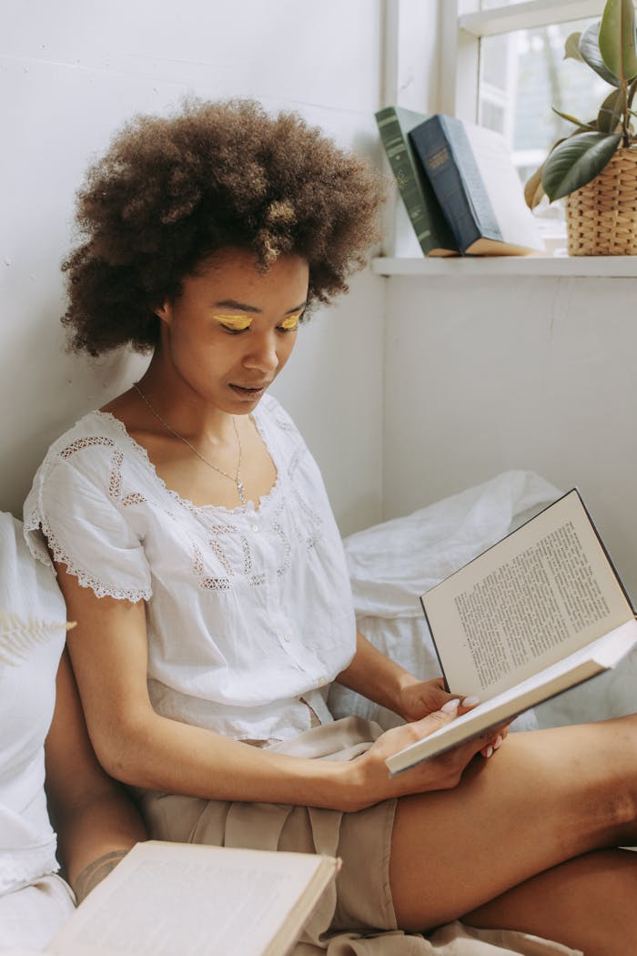 gallery-1 A young woman with natural hair enjoys reading a book by the window with warm sunlight.