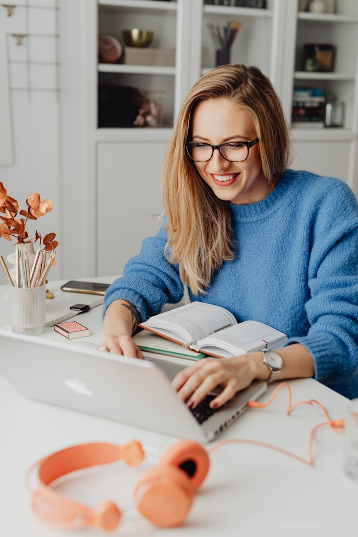 our-services-1 A cheerful woman in a blue sweater working remotely with a laptop in a cozy home setting.