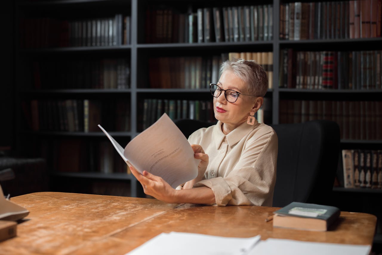 Elegant senior woman reading a manuscript at a wooden desk in a cozy library setting, surrounded by bookshelves.