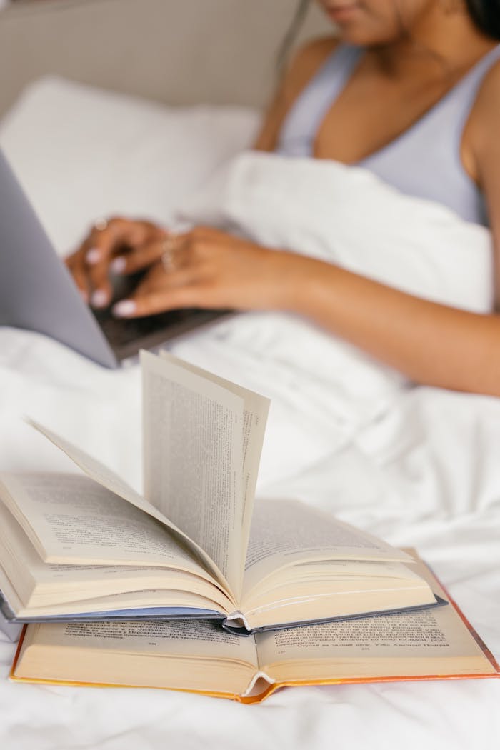 about-01 A person comfortably reading books and using a laptop in bed, draped in a white blanket.